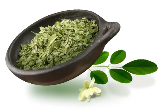 A dark bowl filled with dried green leaves. Next to the bowl, two fresh green leaves and a small white flower are placed on a reflective surface.