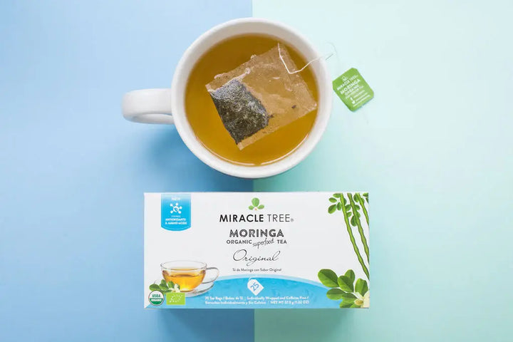 A cup of tea with a teabag on a light blue surface, next to a box of Miracle Tree Moringa Organic Superfood Tea, Original flavor. The box features green moringa leaves and a small image of a teacup.