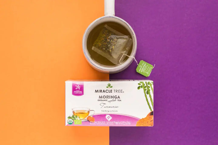 A cup with a tea bag steeping beside a box of Miracle Tree Moringa Organic Superfood Tea in Turmeric flavor. The background is split into orange and purple sections, matching the colors of the tea packaging.