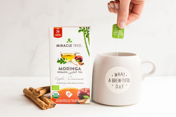 A hand dipping a tea bag into a mug next to an Apple & Cinnamon moringa tea box.