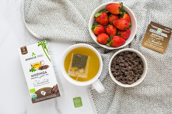 Chocolate moringa tea box beside a cup of tea, strawberries and chocolate pieces.