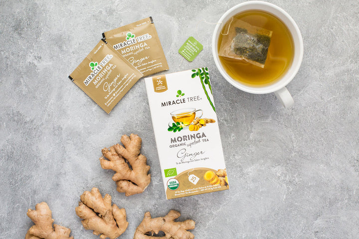 Ginger moringa tea box with sachets, fresh ginger root and a cup of tea.