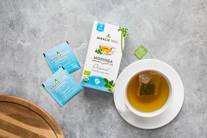 Original moringa tea box with sachets and a cup of tea on a grey counter.