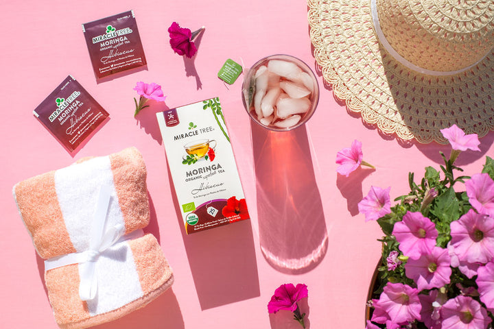 Hibiscus moringa tea box with an iced tea and flowers on a pink table.